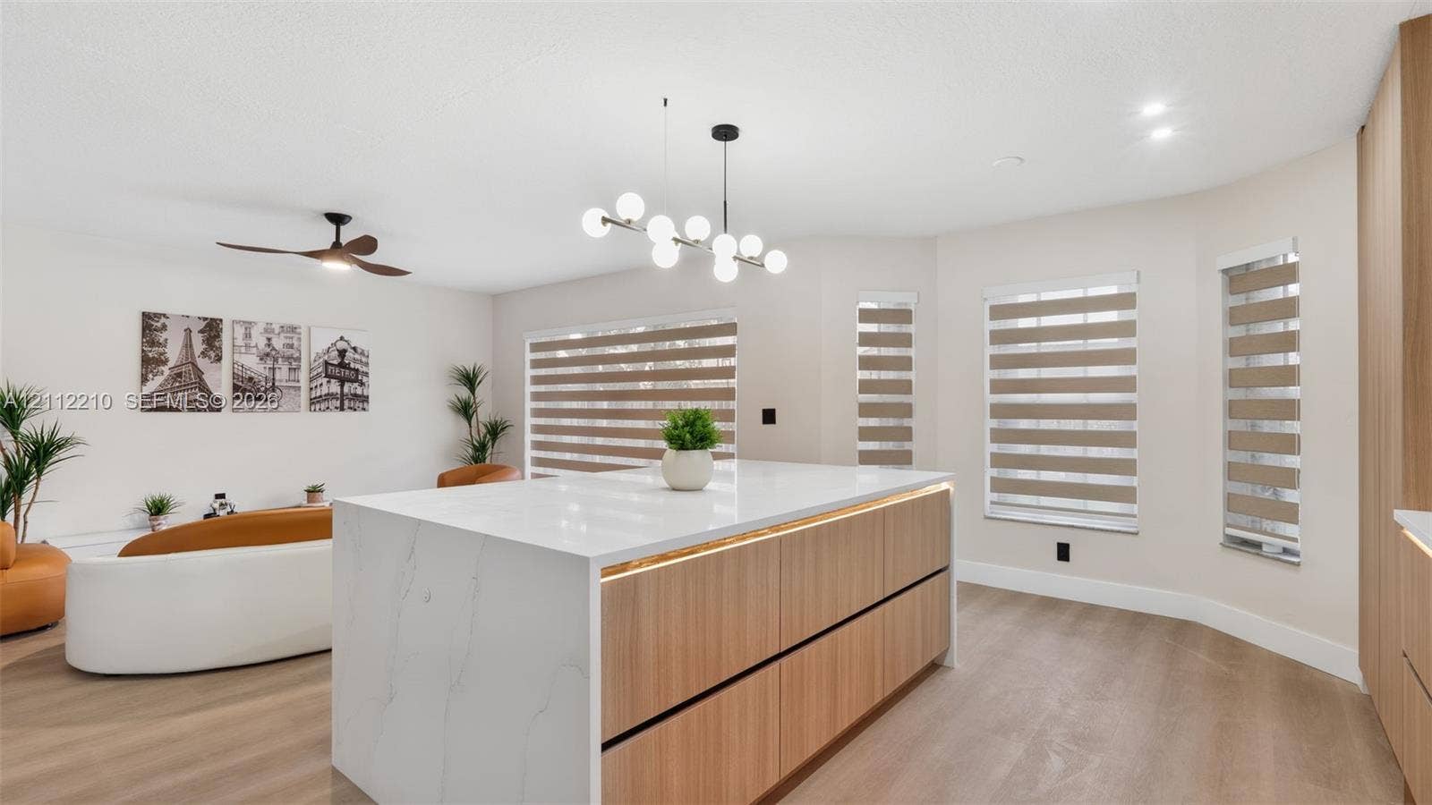 Center island with a stone-finish countertop and integrated wood-finish cabinetry, illuminated by a multi-light contemporary chandelier, featuring light-toned wood-finish flooring and dual-tone window shades