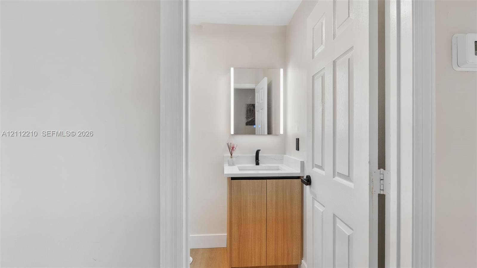 Light-toned wood vanity with a white countertop, integrated rectangular sink, matte black faucet, and rectangular LED-backlit mirror