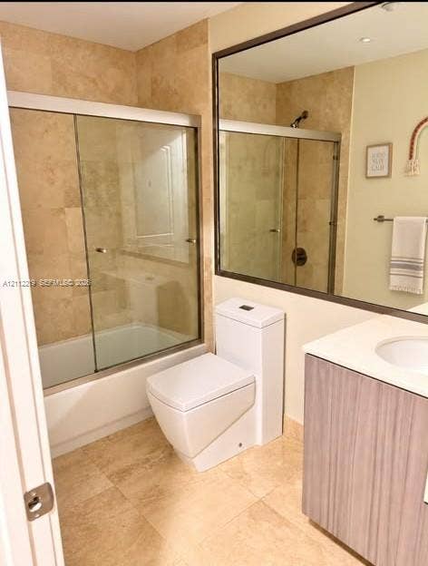 Full bathroom featuring natural stone tiling, a glass-enclosed tub and shower combination, a modern one-piece toilet, a large wall-mounted mirror, and a wood-finish vanity with an integrated basin