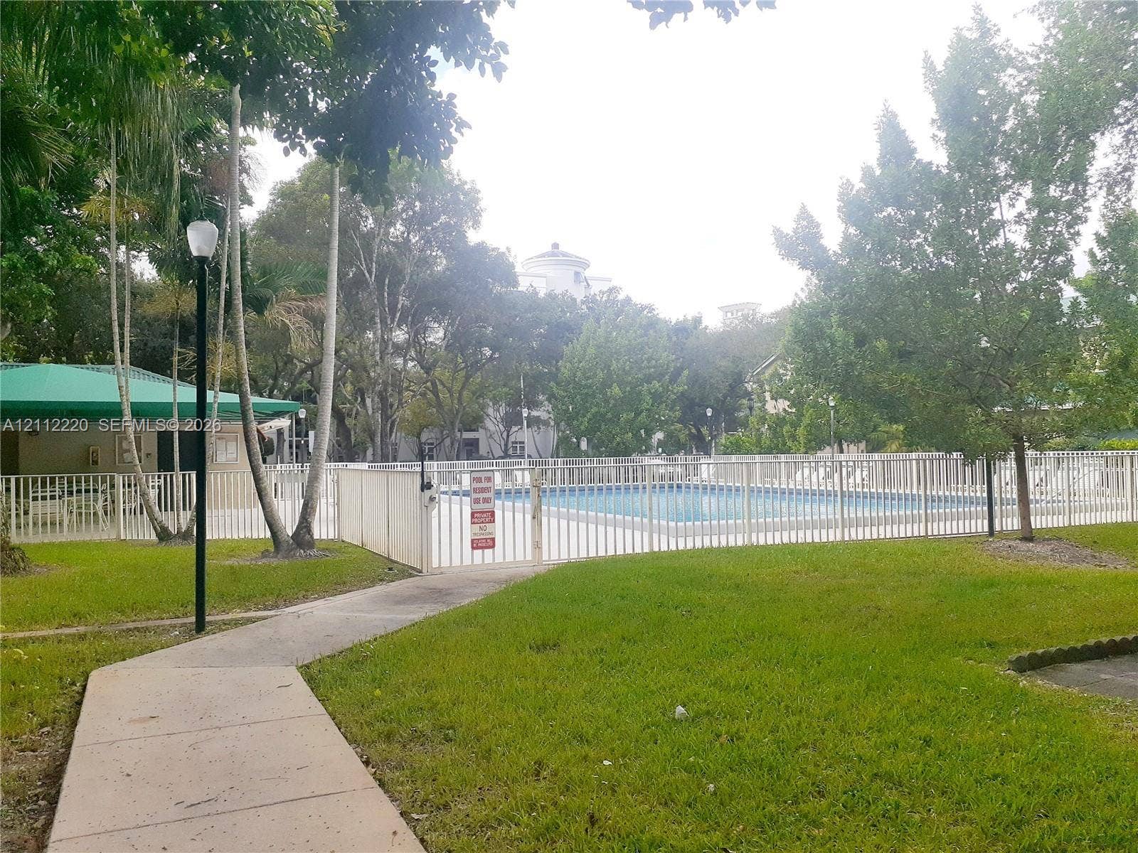 508 W Westree Lane, #508, Plantation, Plantation, FL 33324 Swimming pool enclosed by white metal fencing, featuring an integrated cabana with a green canopy, concrete walkway, and mature landscaping