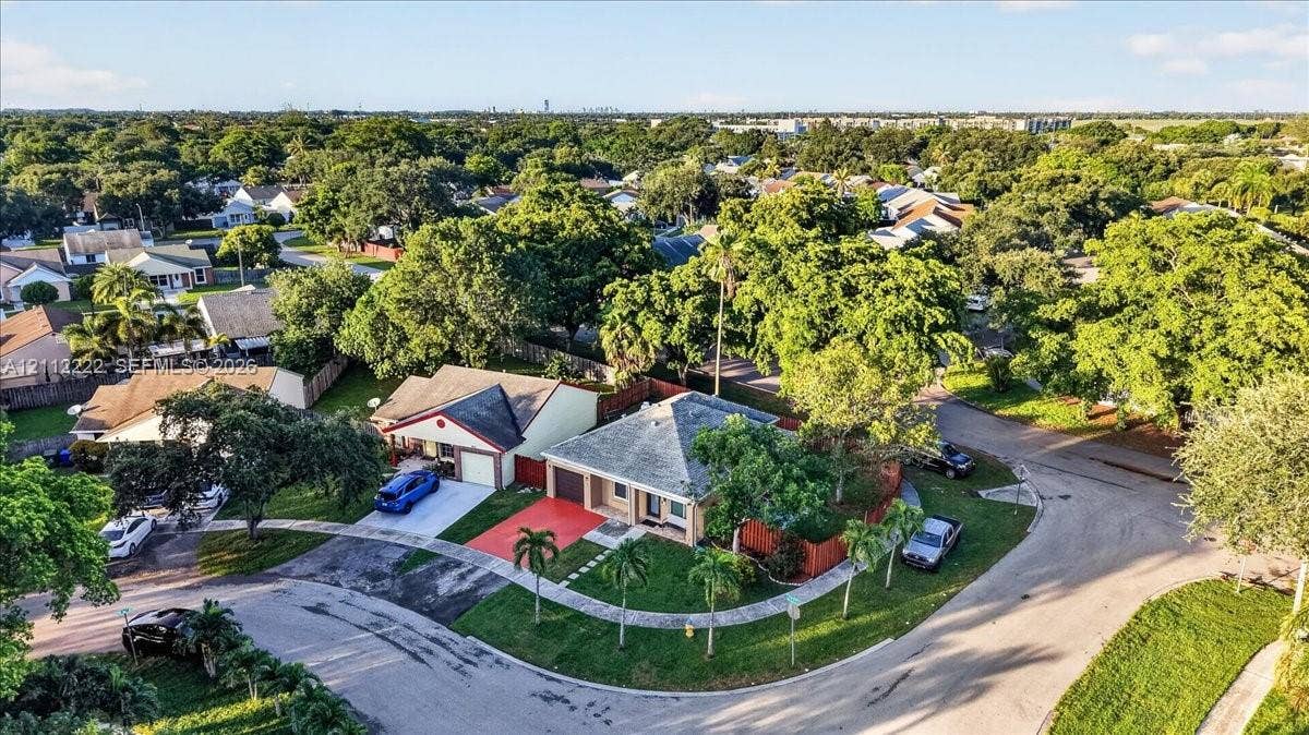 Aerial perspective of a corner lot residence featuring a hipped roof, attached garage, paved driveway, and mature landscaping