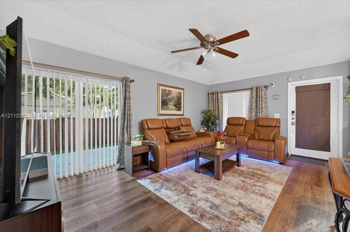 Living area featuring wood-finish flooring, a tray ceiling with a multi-blade ceiling fan, and sliding glass doors with vertical blinds providing exterior access