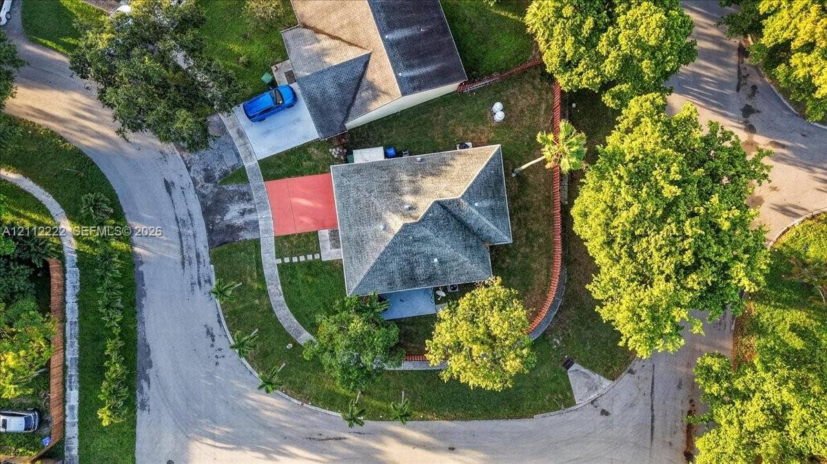 Aerial perspective of a corner lot property featuring a hip roof design, concrete driveway, paved walkway, and mature landscape elements