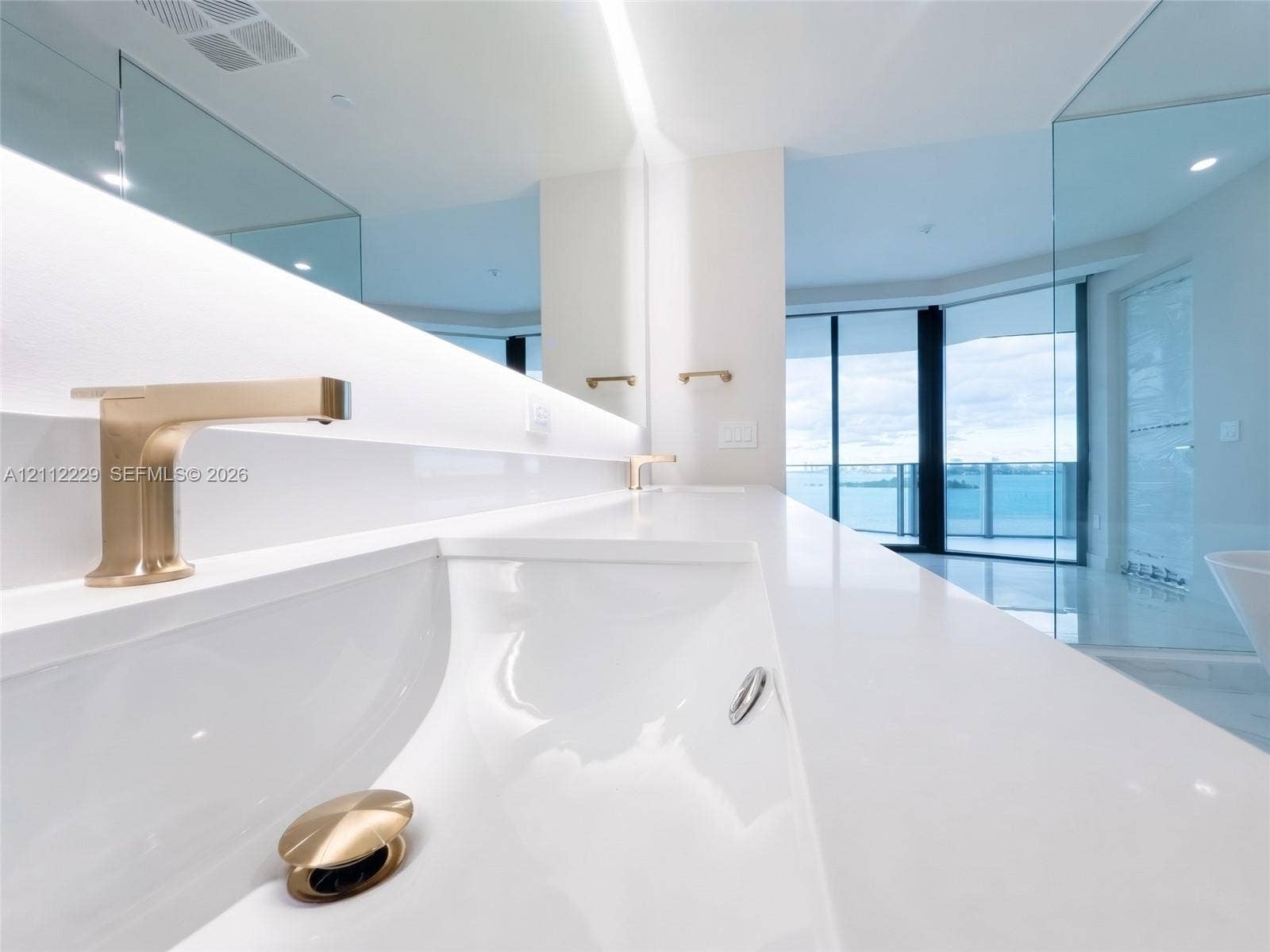 725 NE 24th Street, #706, Miami, Miami, FL 33137 Minimalist vanity featuring a seamless white solid-surface countertop, integrated basin, brushed gold wall-mounted faucets, and an illuminated mirror