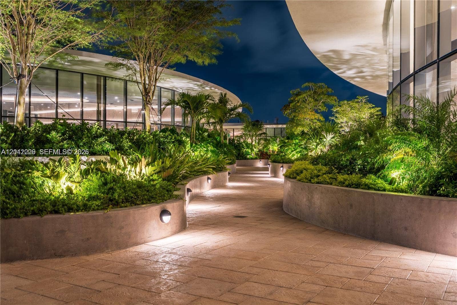 725 NE 24th Street, #706, Miami, Miami, FL 33137 Curved exterior walkway featuring stone-tiled paving, tiered concrete retaining walls, integrated low-level lighting, and floor-to-ceiling glass panel walls
