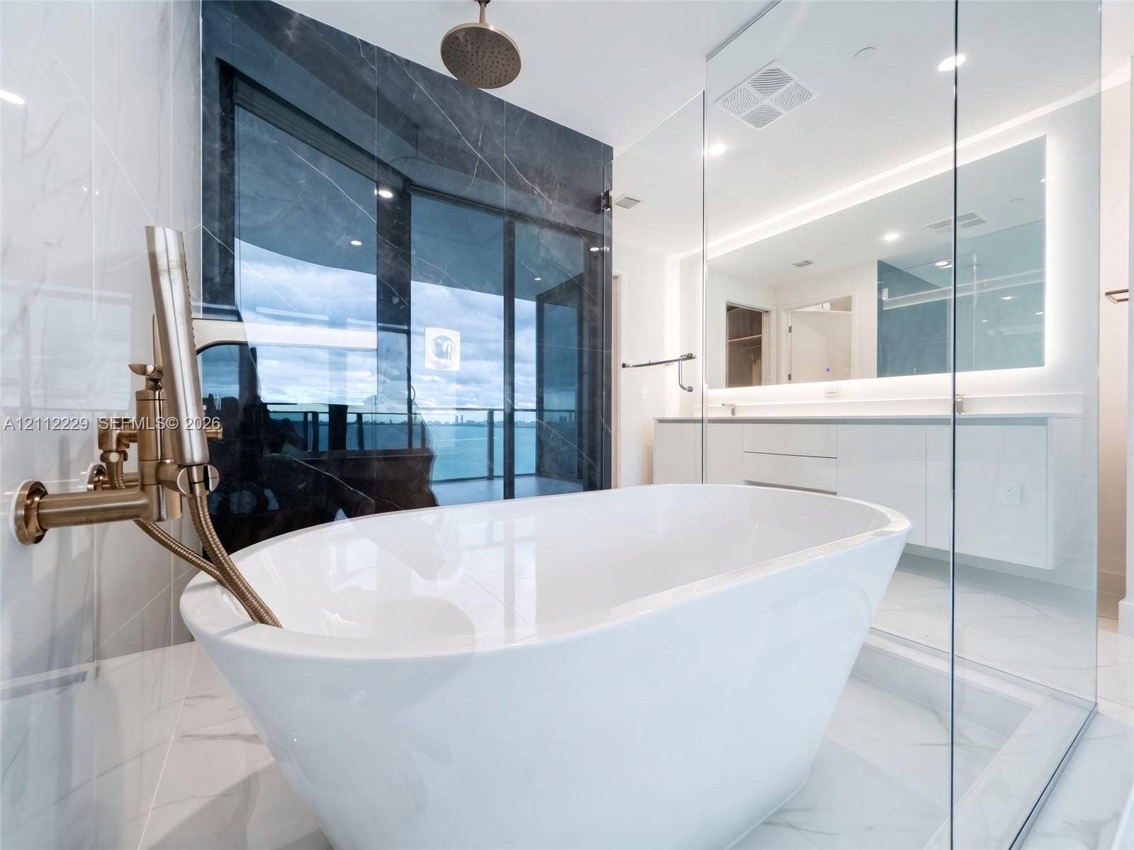 725 NE 24th Street, #706, Miami, Miami, FL 33137 Freestanding soaking tub with brushed gold hardware, glass-enclosed shower, dual-vanity cabinetry, backlit wall mirror, and large-format tile flooring