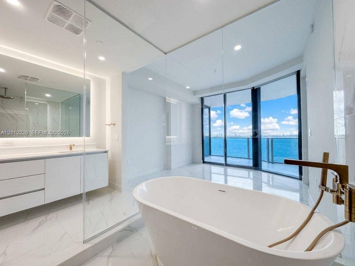 725 NE 24th Street, #706, Miami, Miami, FL 33137 Freestanding soaking tub with a metallic faucet, white cabinetry featuring a light-toned countertop, glass-enclosed shower, large-format stone-finish floor tiling, and floor-to-ceiling glass sliding doors opening to a balcony