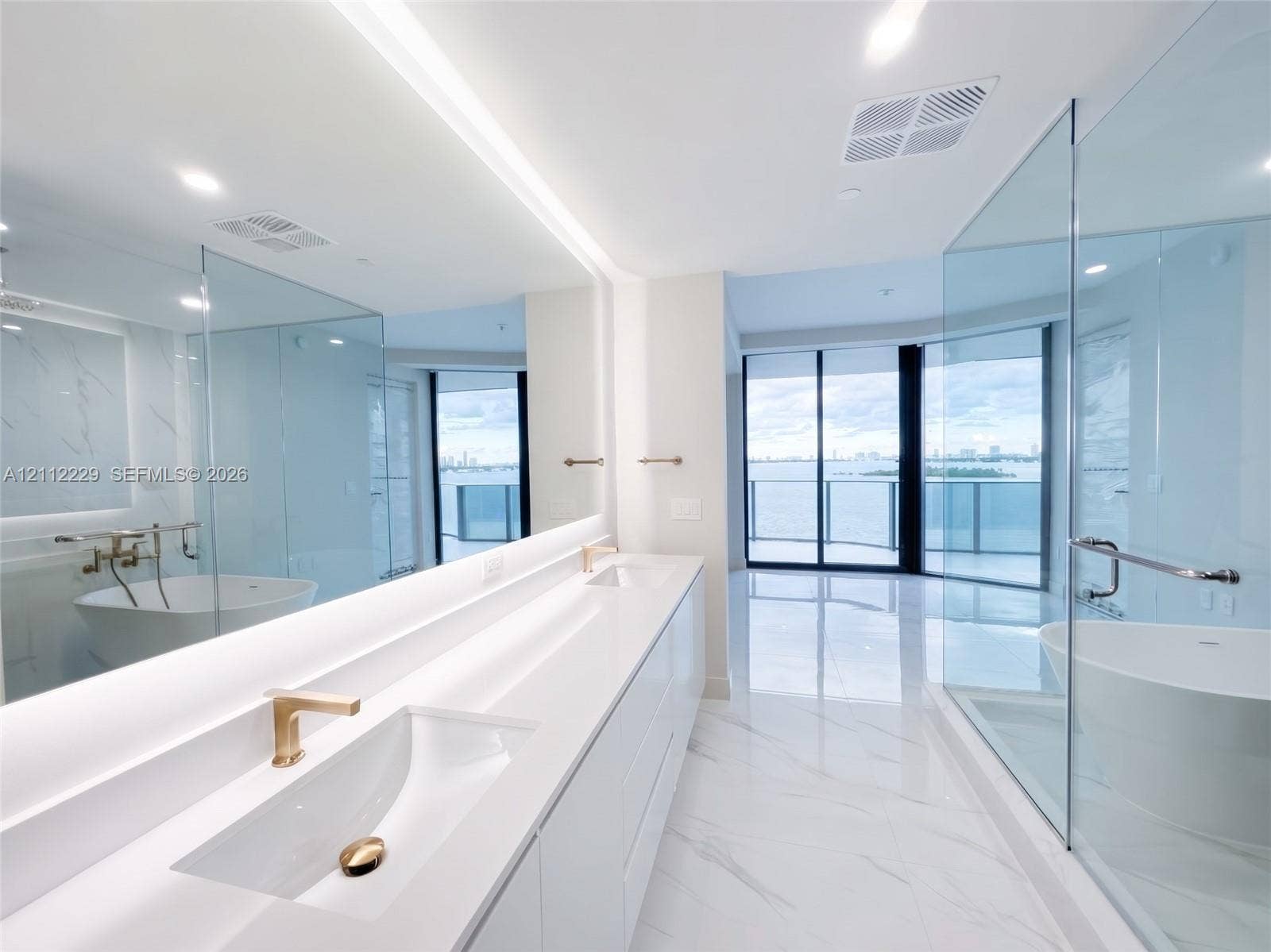 725 NE 24th Street, #706, Miami, Miami, FL 33137 Dual-sink vanity with white countertops and gold-toned fixtures, floor-to-ceiling glass-enclosed shower, freestanding soaking tub, expansive wall-to-wall mirror with integrated lighting, and polished stone-finish flooring