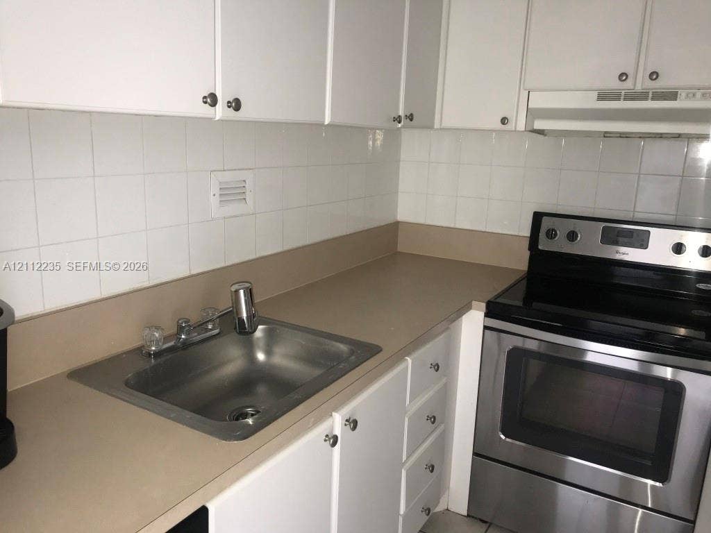 Kitchen featuring white cabinetry, solid surface countertops, stainless steel electric range, square-tile backsplash, and drop-in sink with dual-handle faucet