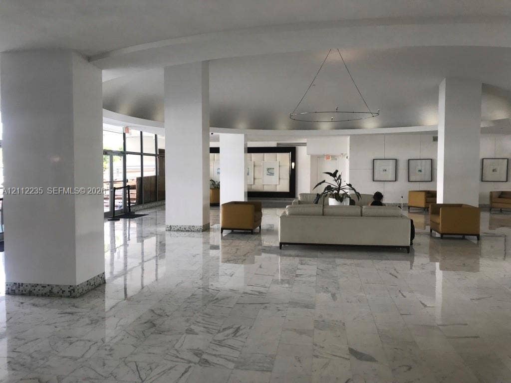 Spacious lobby featuring polished stone flooring, circular recessed ceiling detail, structural support pillars with speckled trim, and floor-to-ceiling glass paneling