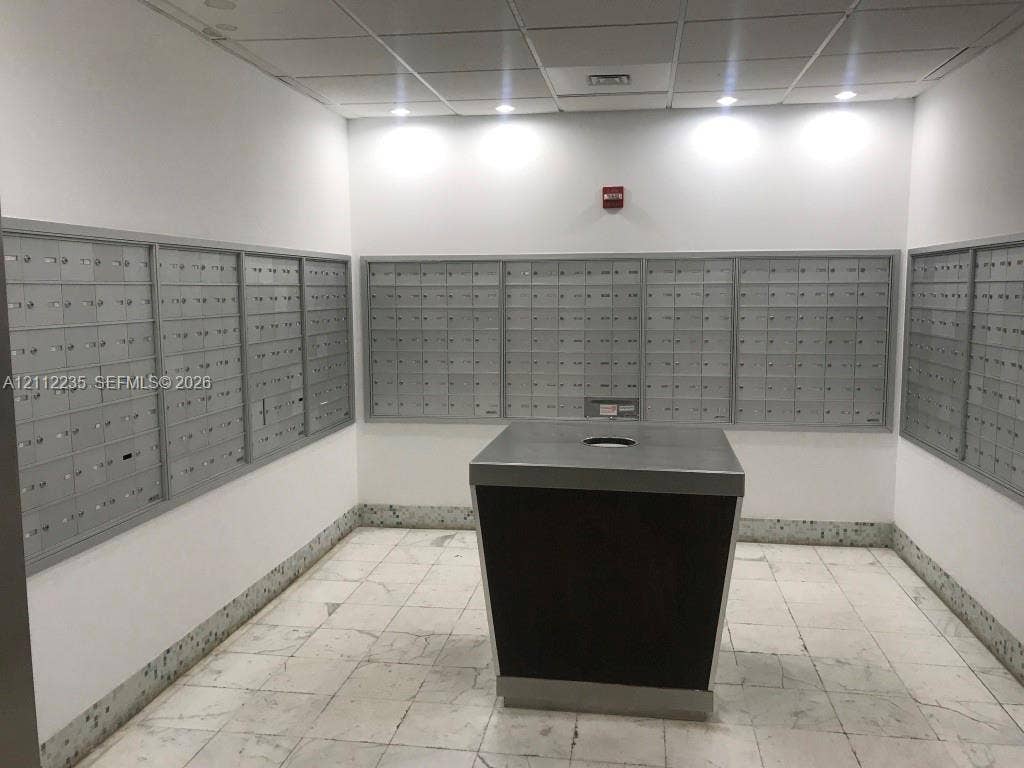 Mail room featuring multiple banks of wall-mounted metal mailboxes, central parcel station with stone countertop, recessed lighting, and stone flooring