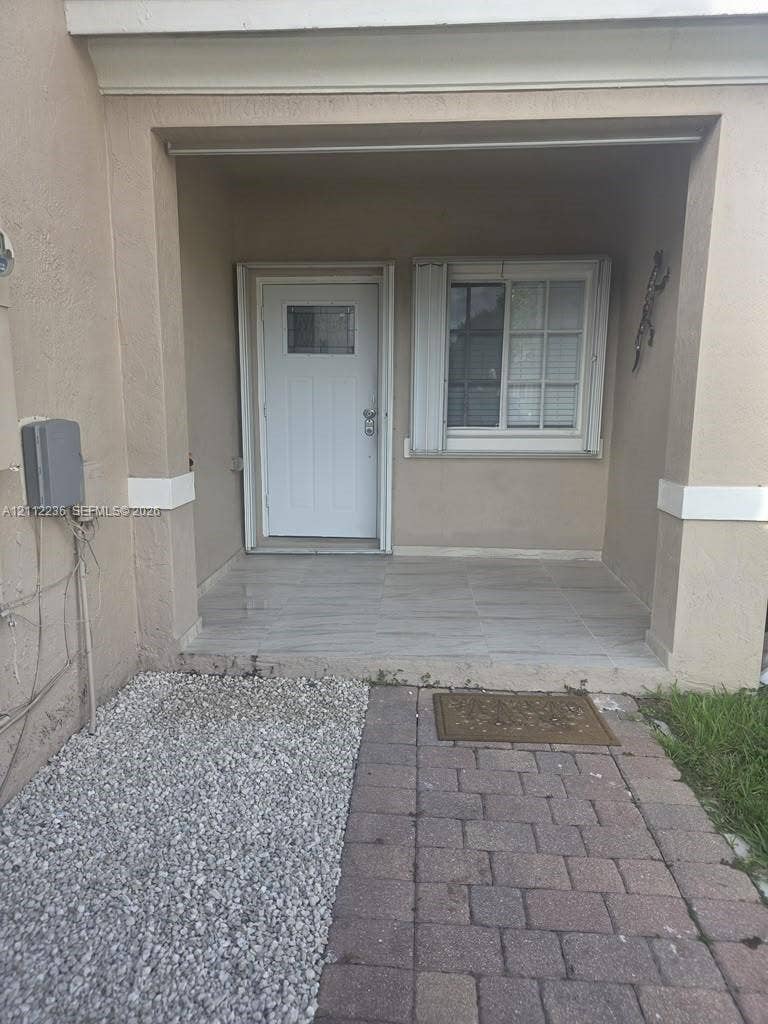 13946 SW 276th Street, #0, Homestead, FL 33032 Covered entryway featuring a stone-tile porch, a white entry door with an integrated window, a sliding window with shutters, and a paver stone walkway