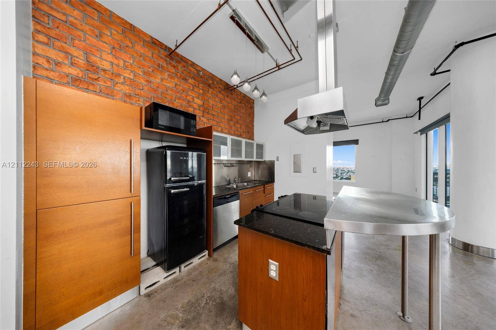 Kitchen featuring exposed brick wall, polished concrete flooring, stainless steel island with built-in cooktop, ceiling-mounted range hood, and wood-finish cabinetry
