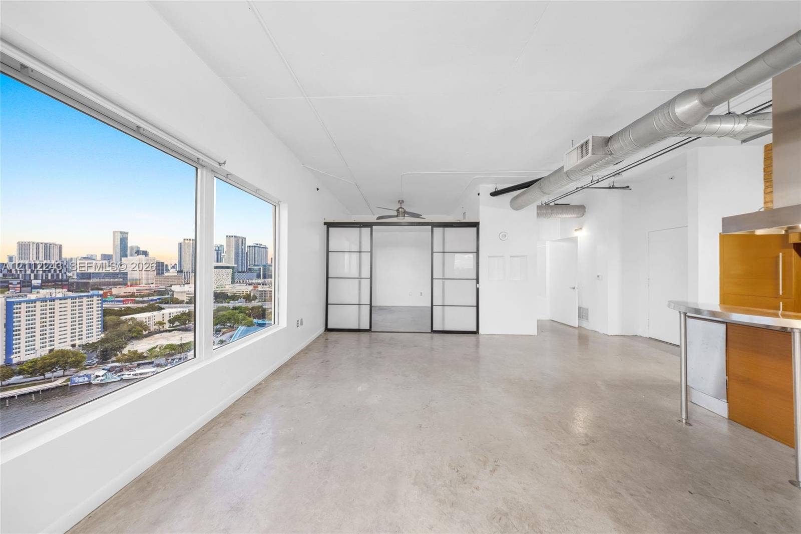 Floor-to-ceiling windows, polished concrete flooring, sliding frosted-glass doors with black metal frames, exposed industrial ductwork, and stainless steel kitchen fixtures