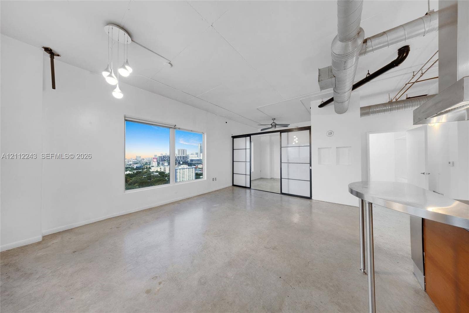Industrial-style loft featuring polished concrete flooring, exposed ductwork, sliding glass-paneled room dividers, a stainless steel breakfast bar, and multi-light pendant fixture