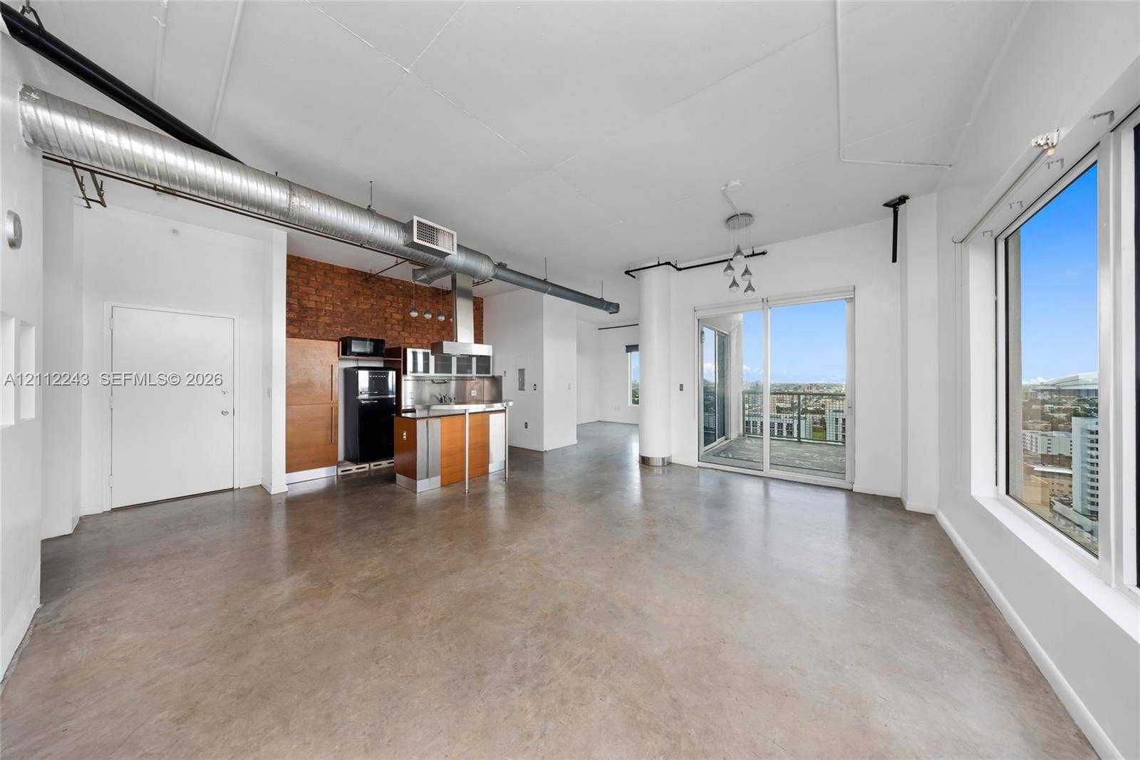 Open-concept interior featuring exposed brick wall accents, polished concrete flooring, industrial-style exposed ductwork, floor-to-ceiling windows, and access to a private balcony
