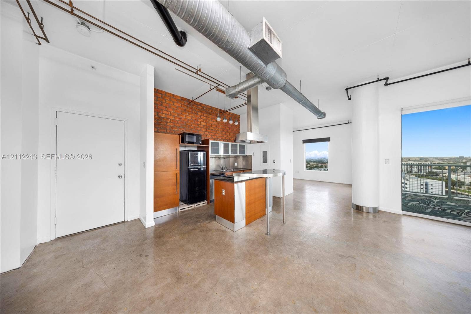 Industrial-style interior featuring polished concrete flooring, exposed brick accent wall, exposed ductwork, and floor-to-ceiling glass door providing exterior access