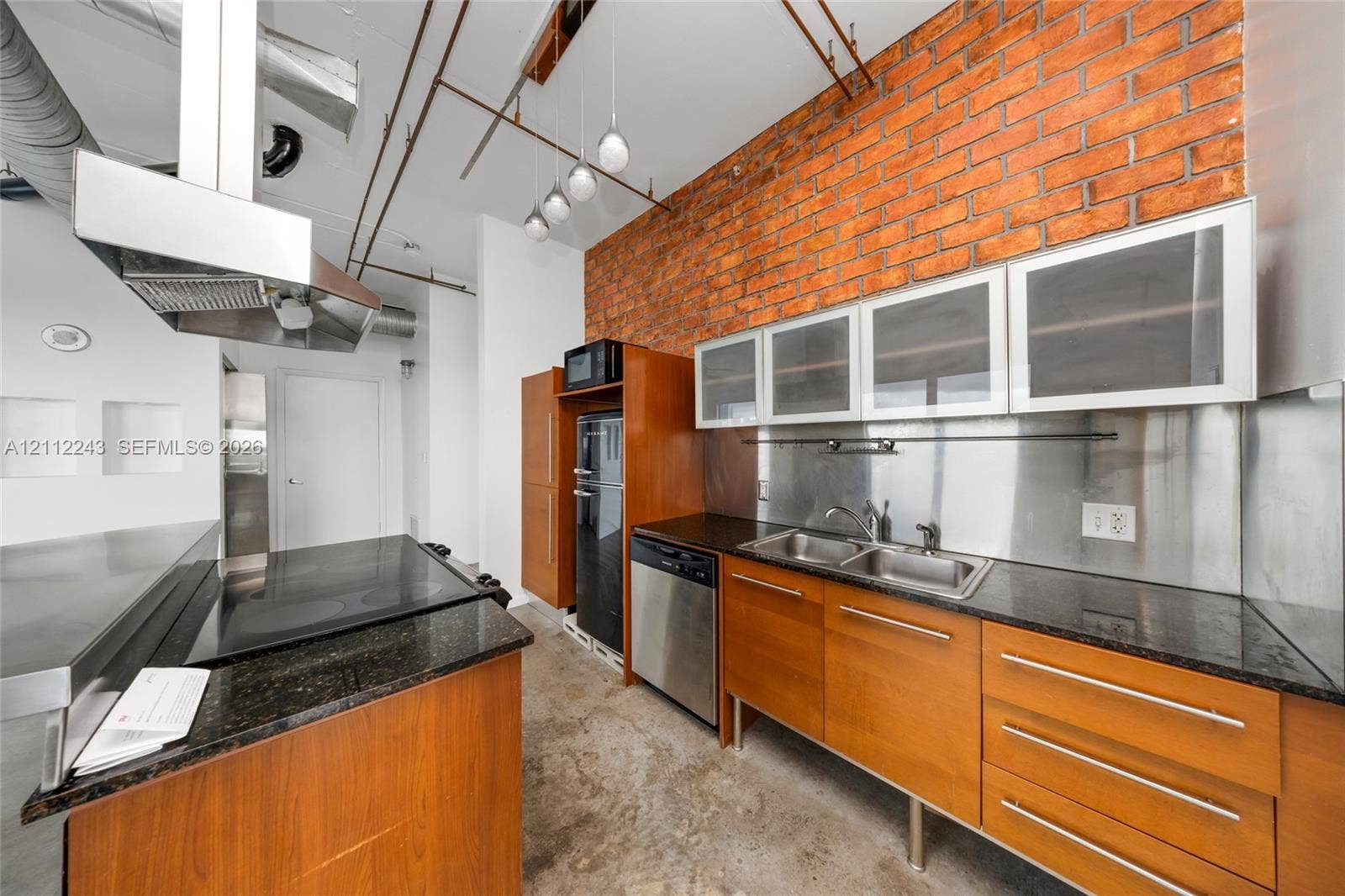 Kitchen featuring an exposed brick accent wall, stone-finish countertops, wood-finish cabinetry, stainless steel backsplash, and built-in stainless steel appliances