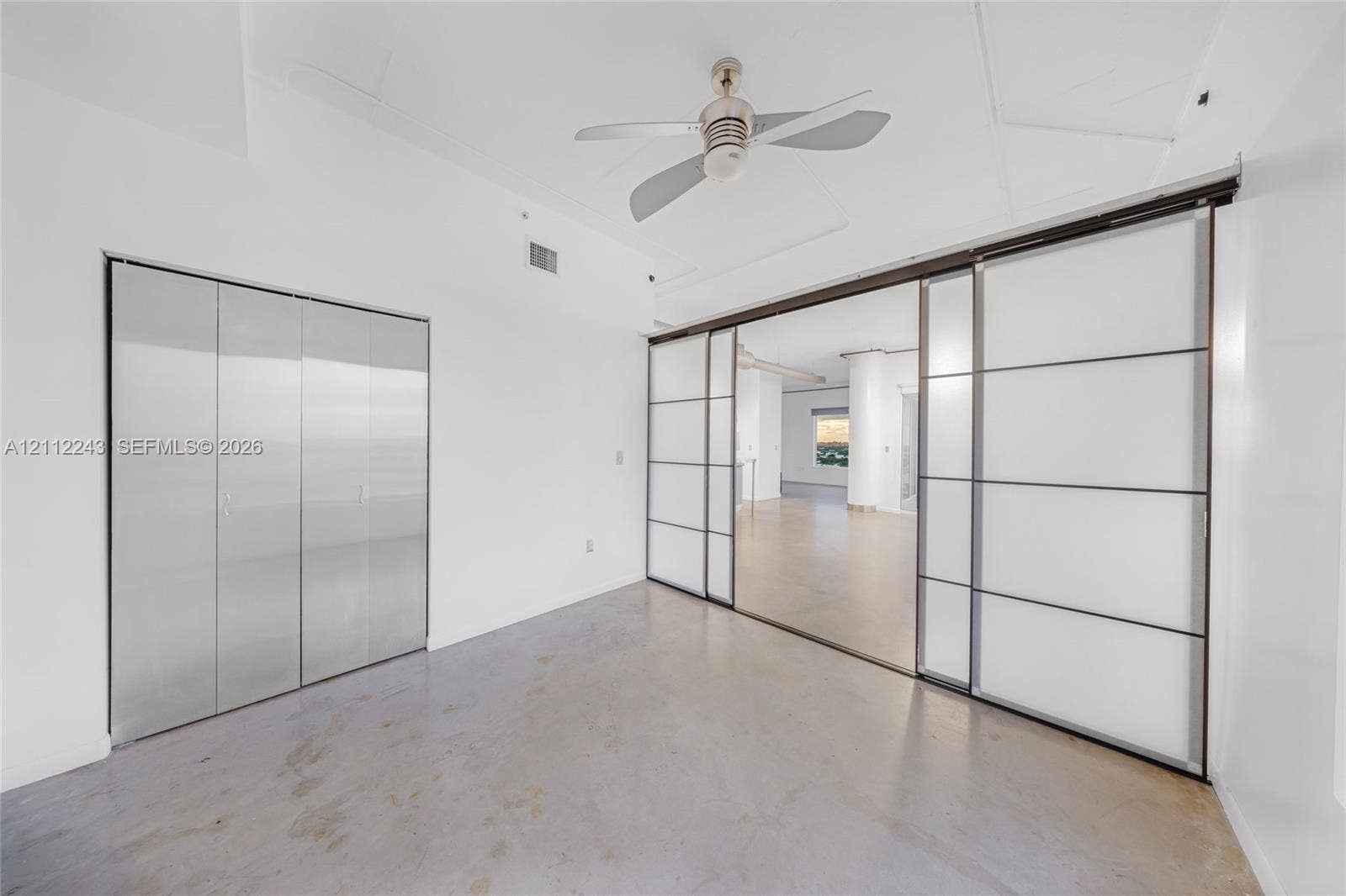Interior space featuring polished concrete flooring, metallic folding closet doors, large frosted glass sliding panels with dark frames, and a ceiling-mounted fan