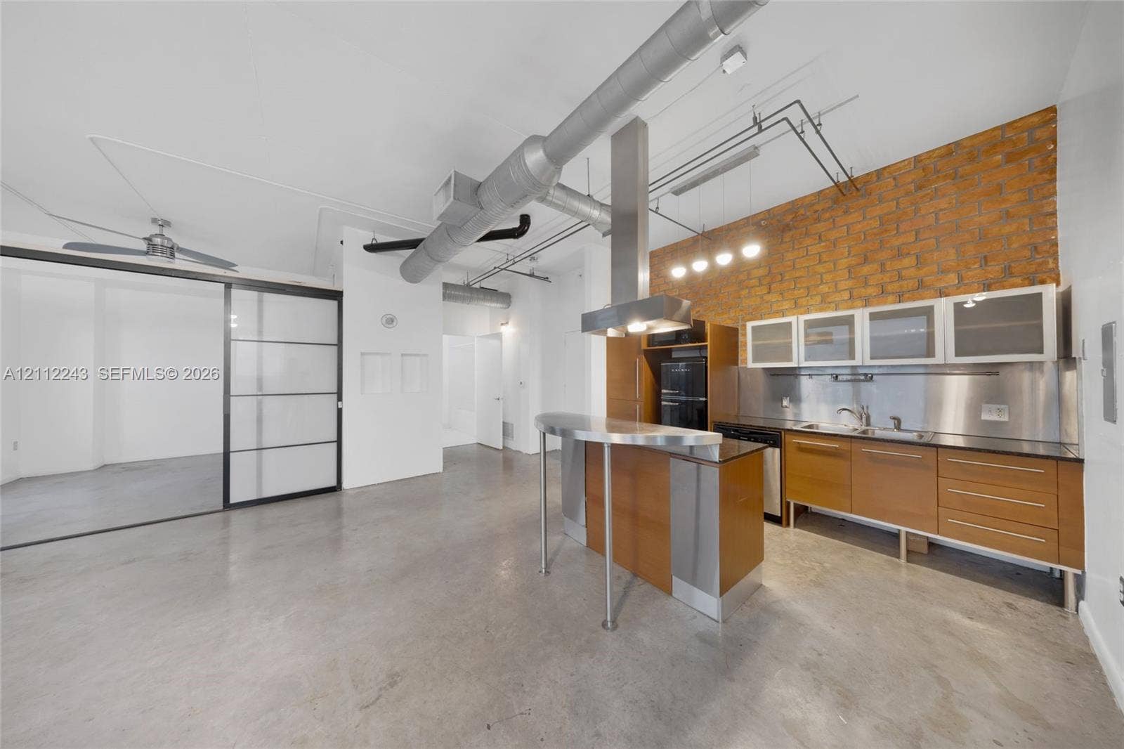 Industrial-style interior featuring exposed brick walls, polished concrete flooring, stainless steel backsplash, custom cabinetry with glass-panel upper storage, and stainless steel kitchen peninsula with integrated venting hood