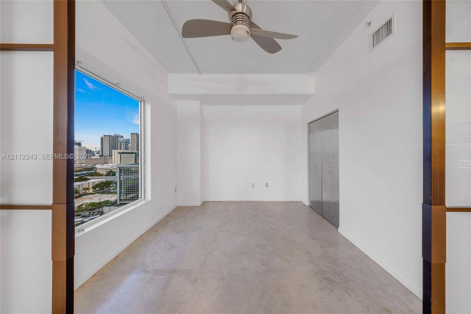 Interior space featuring polished concrete flooring, metallic sliding closet doors, a ceiling-mounted fan with integrated lighting, and a large window showcasing a city skyline