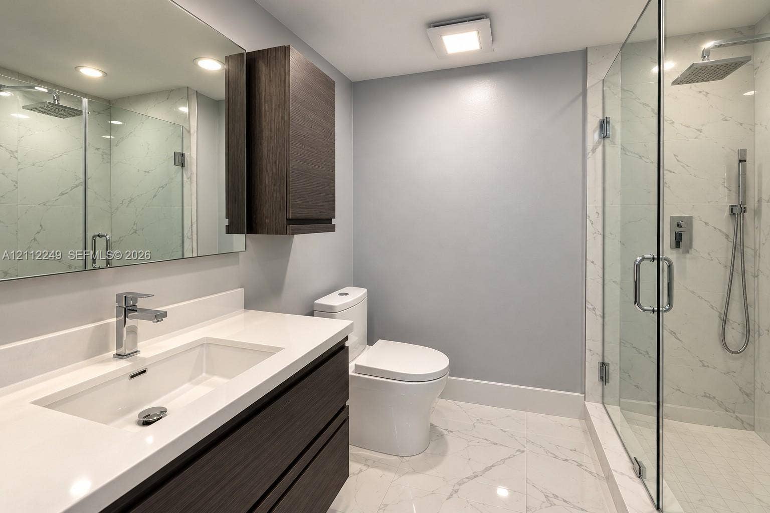 Modern bathroom featuring a dark wood-finish vanity with a white countertop and rectangular undermount sink, large-format tile flooring with marble-patterned veining, a glass-enclosed shower with a rainfall showerhead and handheld sprayer, and a wall-mounted wood-finish storage cabinet