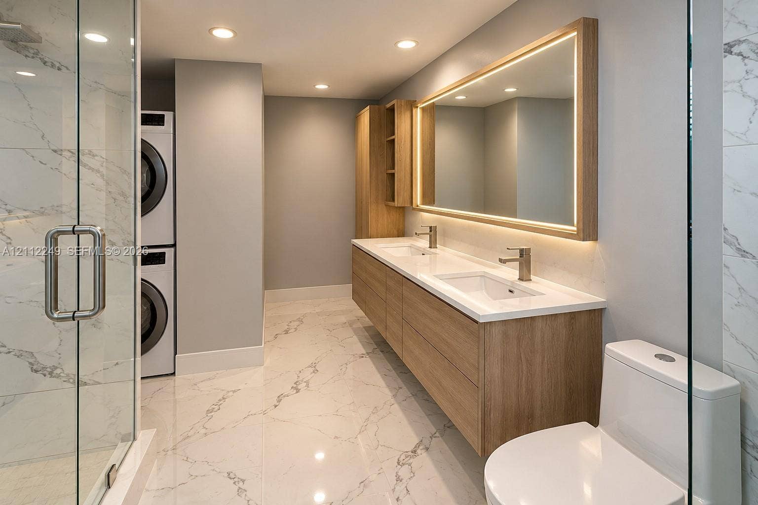 Dual-sink floating vanity with wood-finish cabinetry, integrated LED-backlit mirror, floor-to-ceiling stone-style tile flooring, glass-enclosed shower, and built-in stacked laundry center
