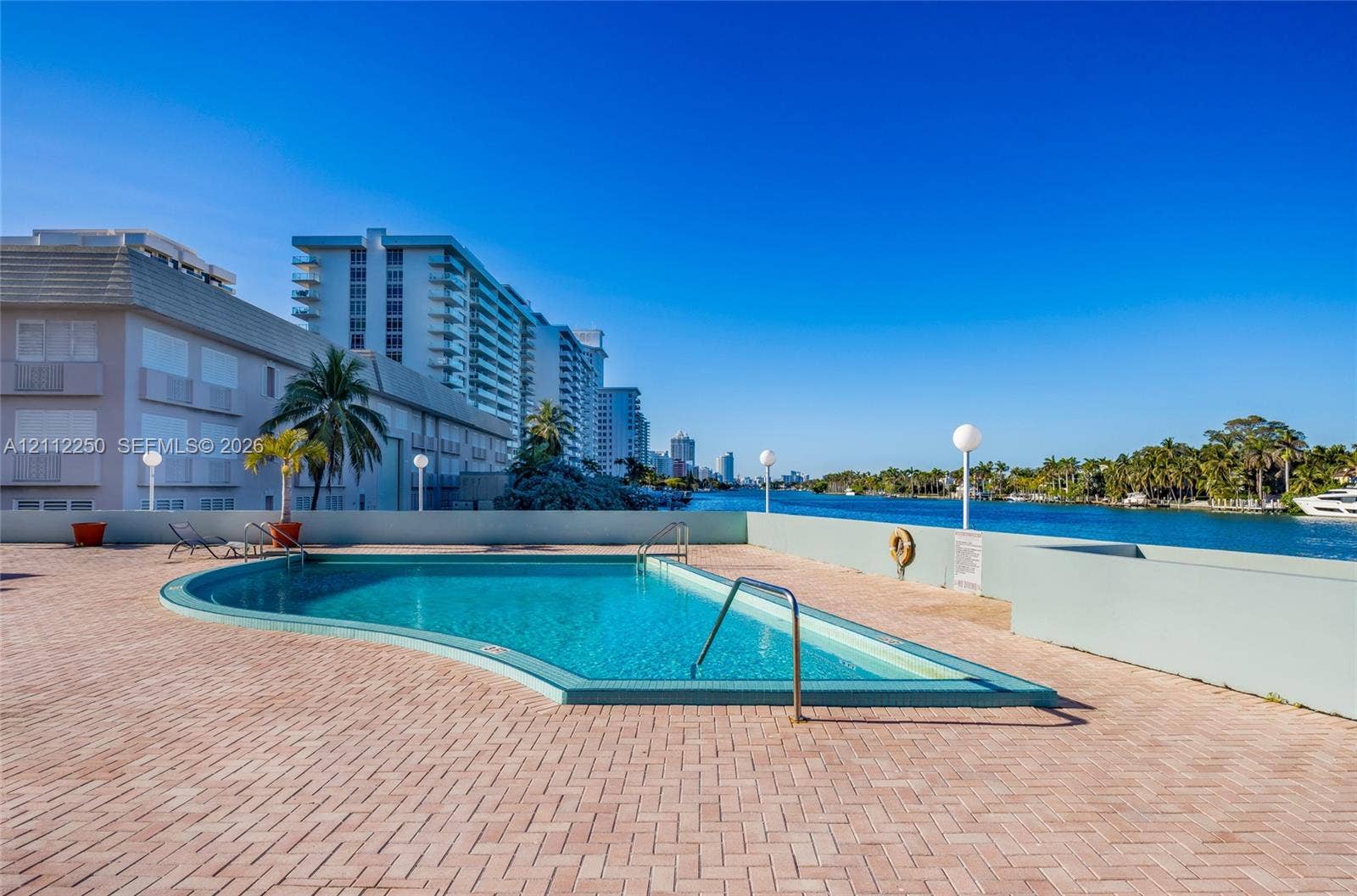 5838 Collins Avenue, #2F, Miami Beach, FL 33140 Curved swimming pool featuring a brick-paved deck, perimeter wall, metal handrails, and direct waterfront positioning