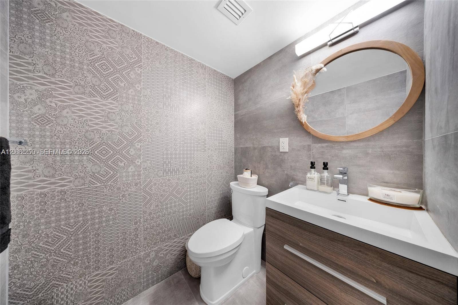 5838 Collins Avenue, #2F, Miami Beach, FL 33140 Powder room featuring a wood-finish vanity with an integrated basin, large-format grey tile walls, a decorative patterned accent wall, and an oval mirror with a natural wood frame