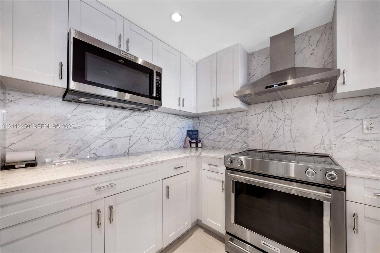 5838 Collins Avenue, #2F, Miami Beach, FL 33140 Kitchen featuring white Shaker-style cabinetry, stone-finish countertops, full-height stone-finish backsplash, stainless steel microwave, and stainless steel range with a matching chimney hood