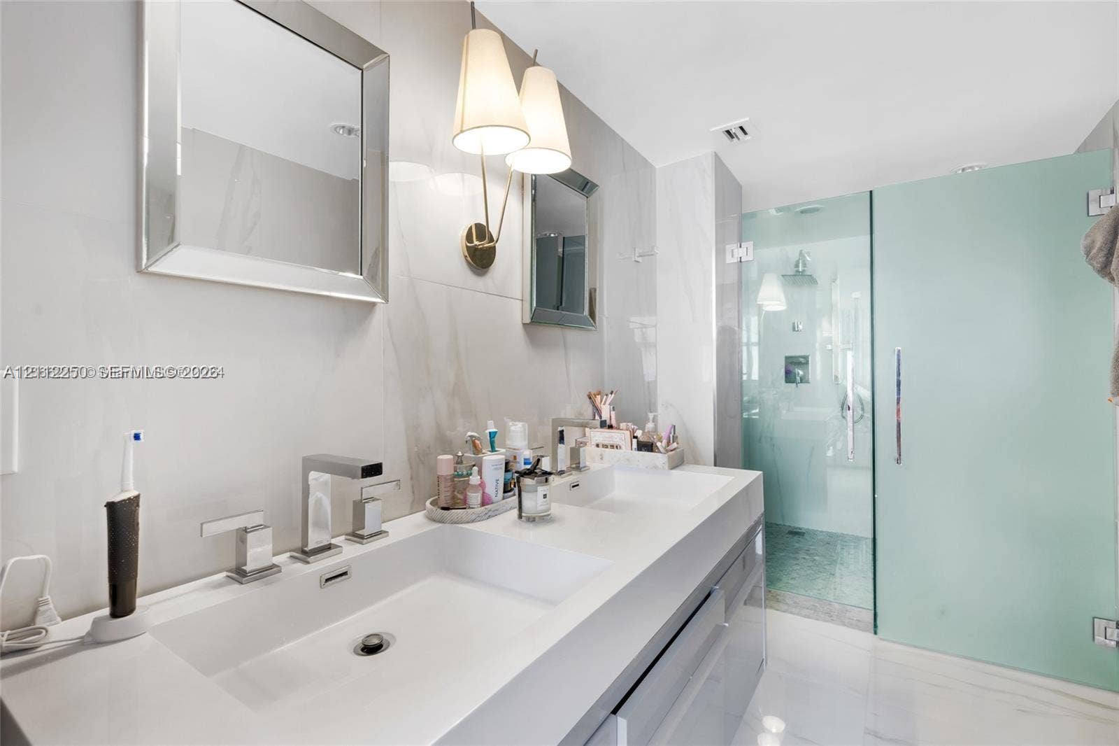 5838 Collins Avenue, #2F, Miami Beach, FL 33140 Dual-sink vanity with a solid-surface countertop, rectangular basins, wall-mounted polished chrome faucets, and frameless glass shower enclosure