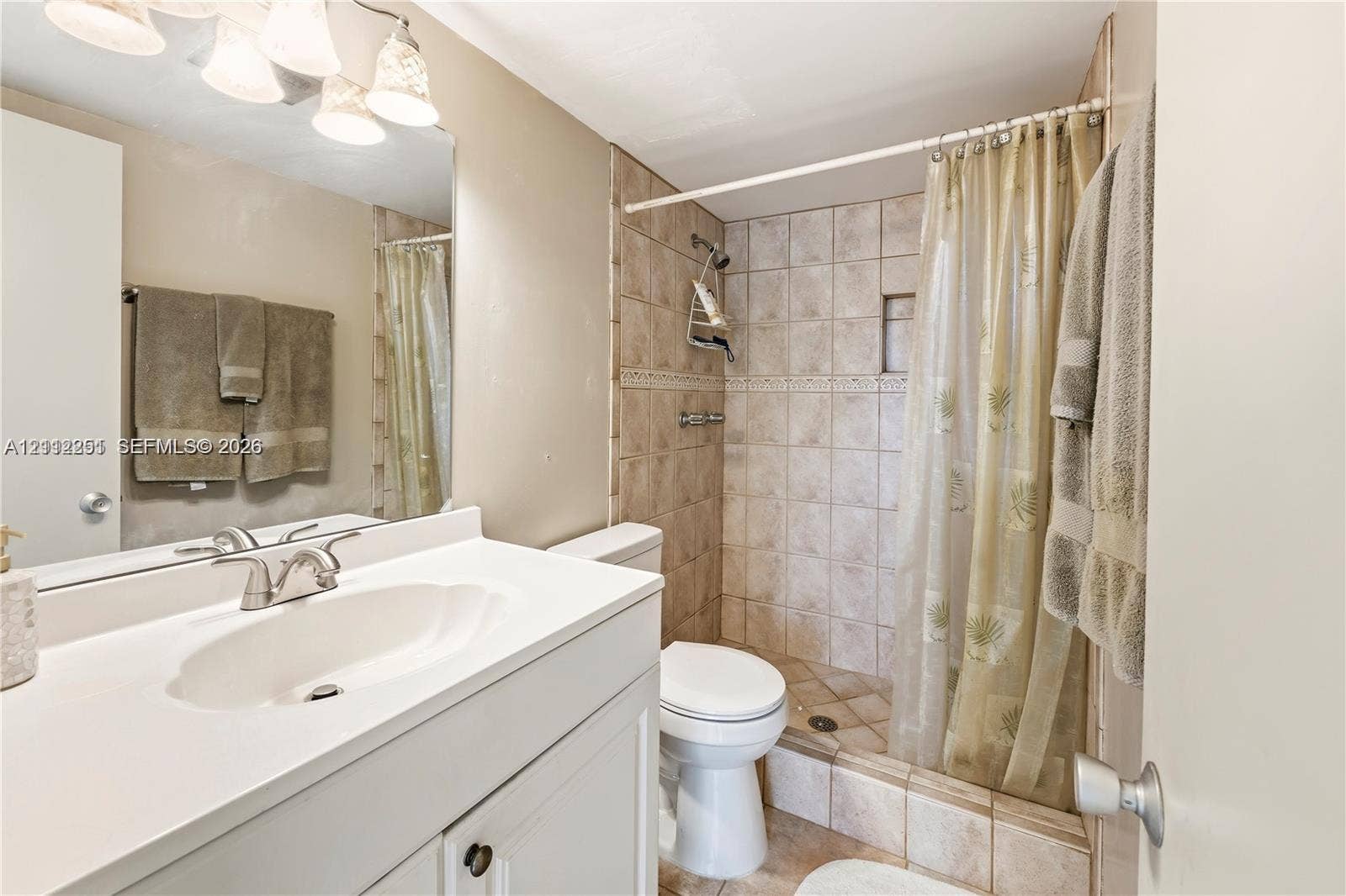 4025 N Federal Highway, #226C, Oakland Park, FL 33308 Bathroom featuring a white vanity with an integrated countertop, polished chrome faucet, full-width mirror, light fixture with four frosted glass shades, and a tiled shower stall with a decorative accent border
