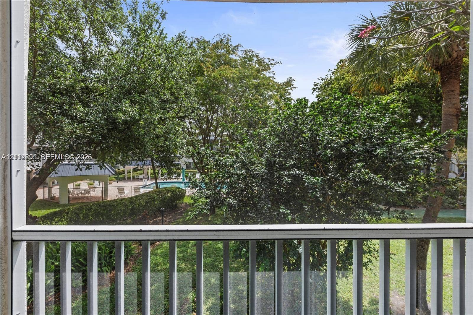 4025 N Federal Highway, #226C, Oakland Park, FL 33308 Screened-in balcony with white metal railings, overlooking a swimming pool area, manicured landscaping, mature trees, and a pavilion structure