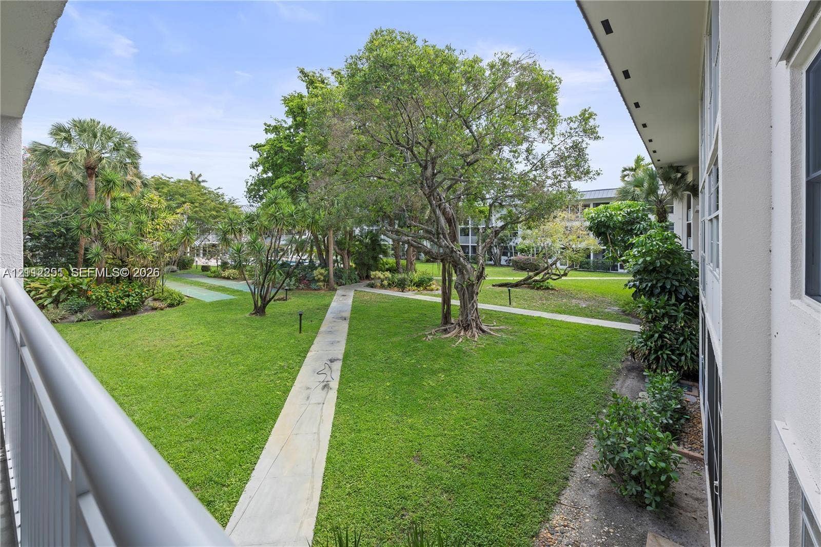 4025 N Federal Highway, #226C, Oakland Park, FL 33308 Covered balcony with white railing overlooking a landscaped common area featuring mature trees, manicured lawn, and concrete walkways