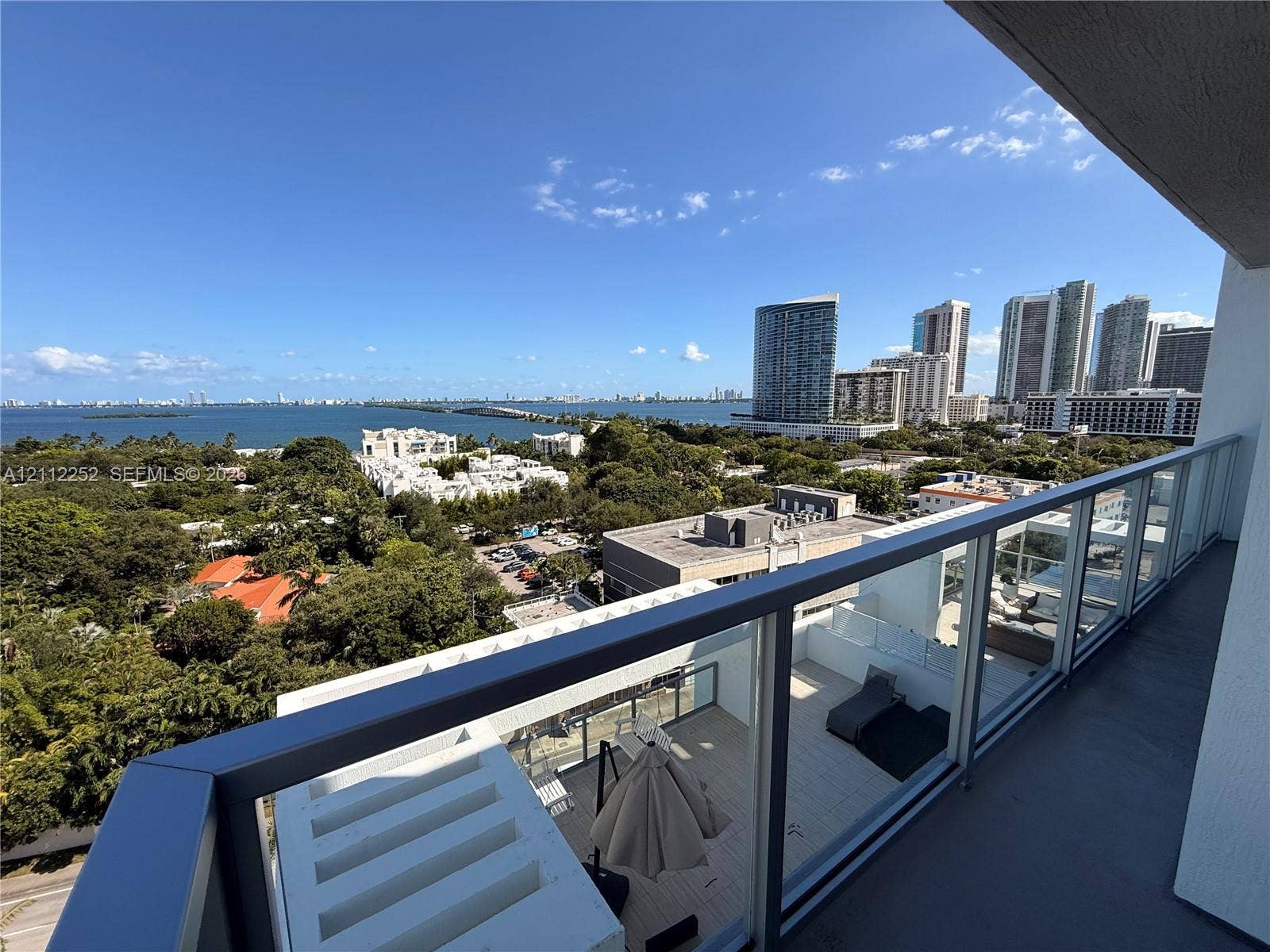 3900 Biscayne Boulevard, #N-1113, Miami, FL 33137 Covered balcony featuring glass railing panels and metal handrails, overlooking a coastal landscape with high-rise structures and open water