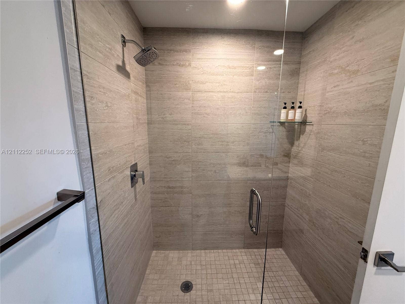 3900 Biscayne Boulevard, #N-1113, Miami, FL 33137 Walk-in shower featuring floor-to-ceiling stone-finish tile surround, frameless glass door, mosaic tile flooring, built-in glass shelving, and chrome hardware