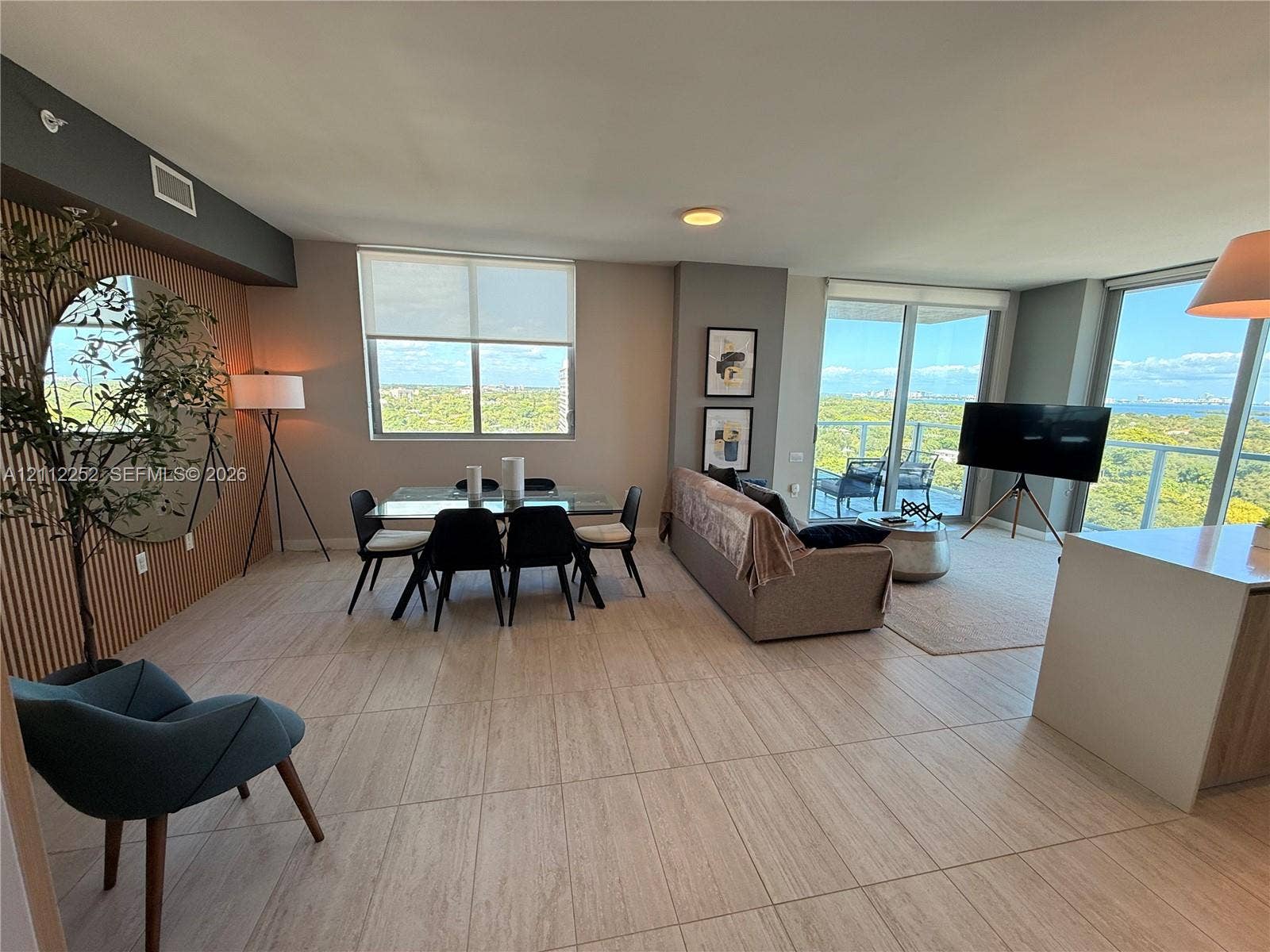3900 Biscayne Boulevard, #N-1113, Miami, FL 33137 Open-concept living area featuring neutral tile flooring, floor-to-ceiling windows, natural wood-slat accent wall, and glass sliding doors providing access to a private balcony