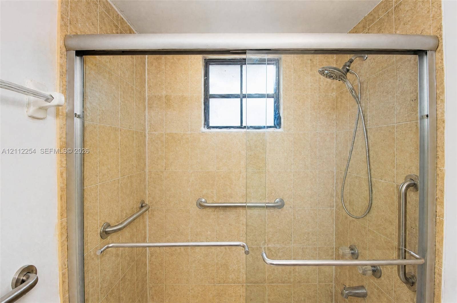 Walk-in shower enclosure featuring tiled wall surrounds, sliding glass doors, multiple stainless steel grab bars, a handheld shower fixture, and an operable window