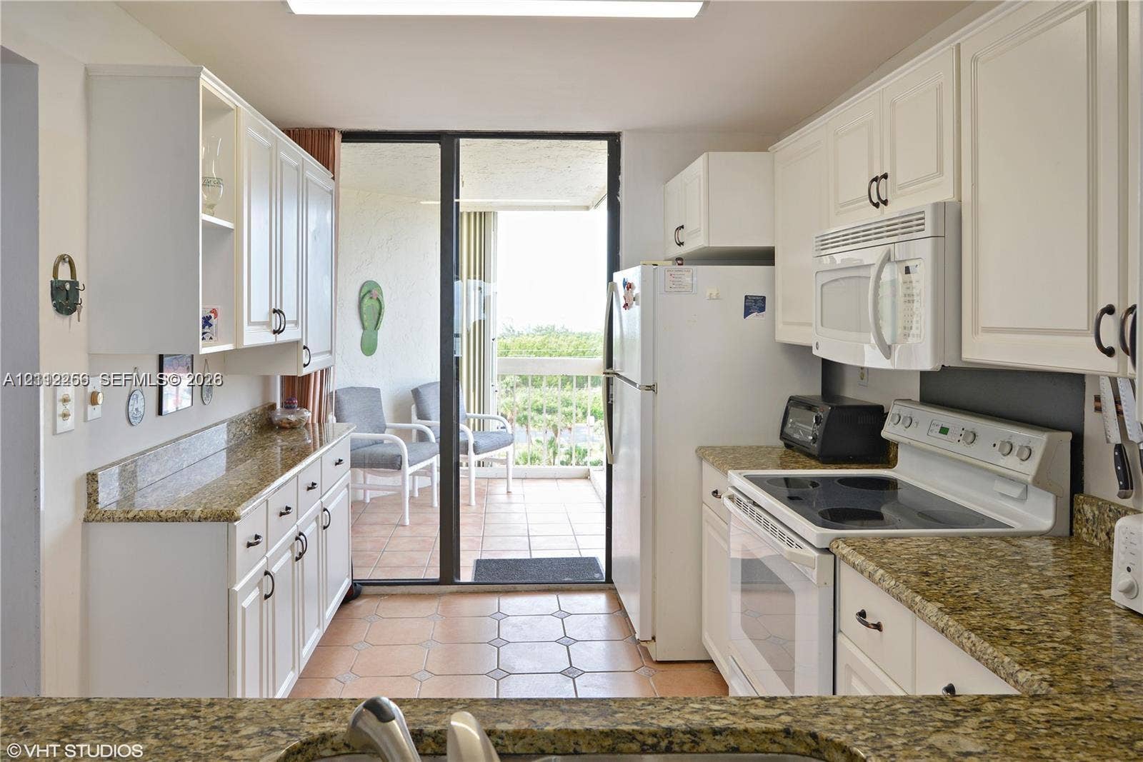 9900 S Ocean Drive, #601, Jensen Beach, FL 34957 Kitchen featuring white shaker-style cabinetry, stone-finish countertops, tile flooring, and integrated white appliances, with sliding glass doors opening to a tiled balcony