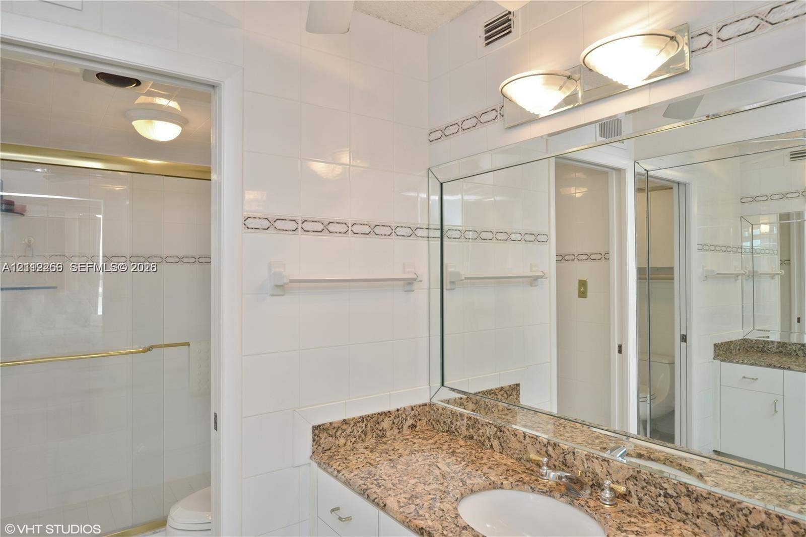 9900 S Ocean Drive, #601, Jensen Beach, FL 34957 Full-width vanity with stone-finish countertop, undermount sink, integrated mirror, glass-enclosed shower with gold-tone hardware, and white wall tiles with decorative accent trim
