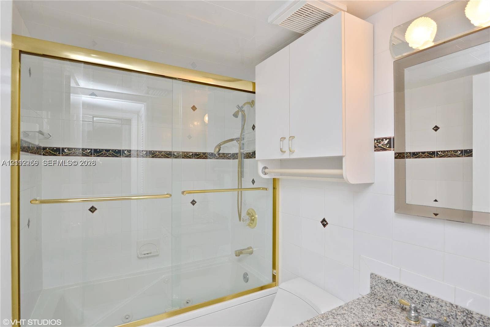 9900 S Ocean Drive, #601, Jensen Beach, FL 34957 Bathroom featuring a tub-shower combination with sliding glass doors, gold-tone metal trim, stone-composite vanity countertop, wall-mounted white cabinetry, and decorative tile accents