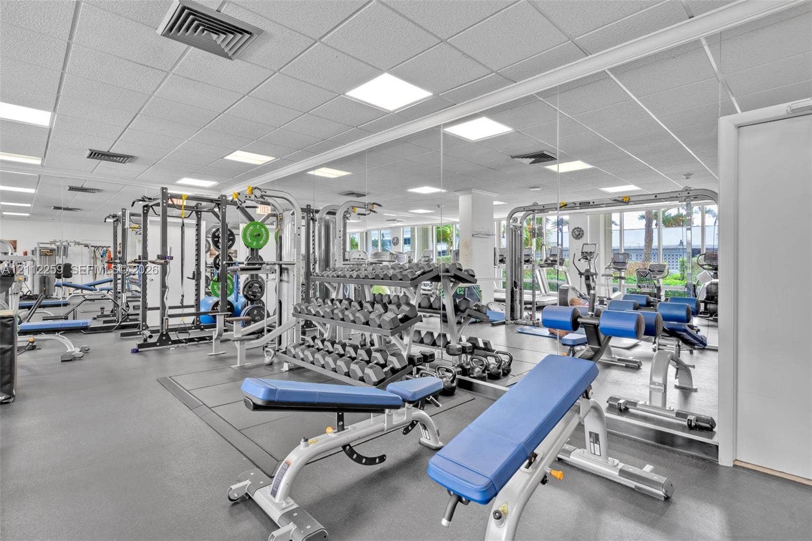 Fitness area featuring a rubberized flooring surface, drop ceiling with recessed panel lighting, and wall-to-wall mirrors