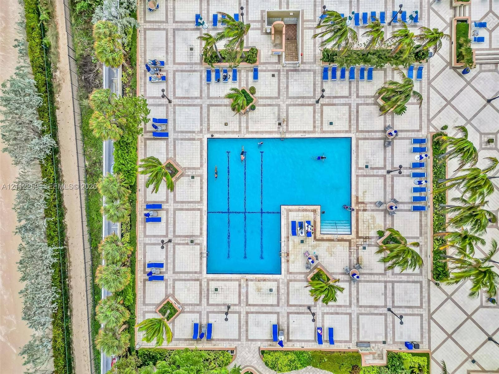 Geometric swimming pool with lap lanes and integrated stairs, surrounded by extensive patterned stone tiling, tropical landscaping, and mature palm trees