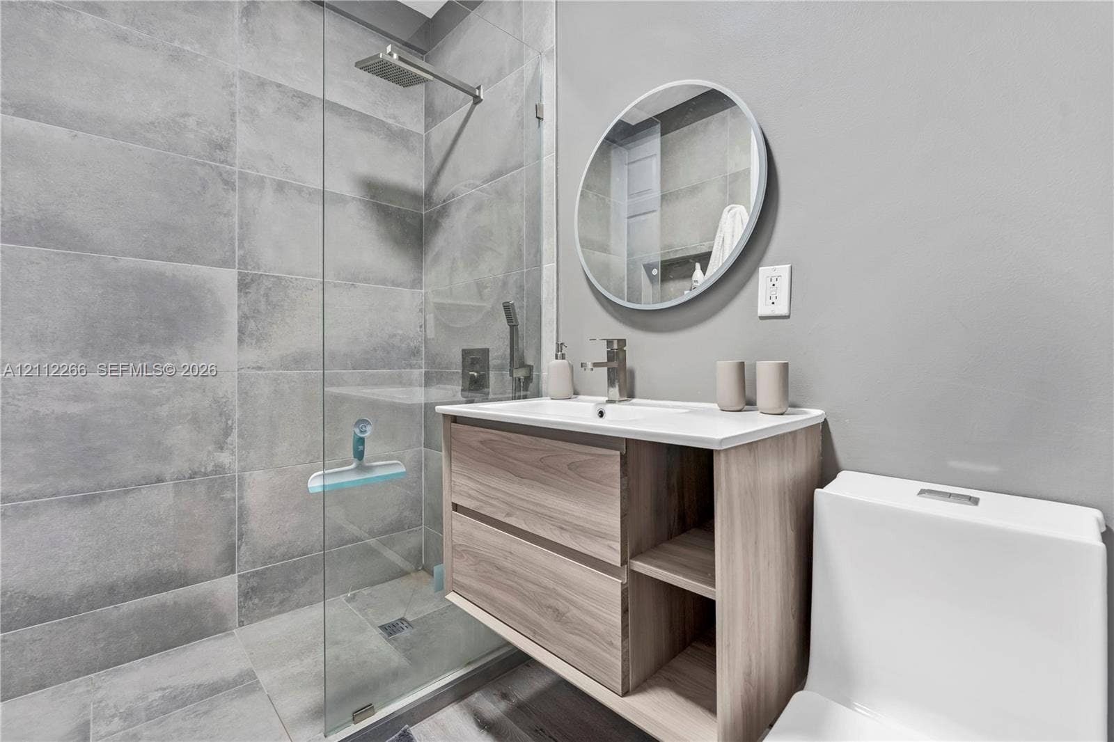 Walk-in shower with large-format grey tile surround, glass partition, and rain-style shower head, featuring a wood-finish floating vanity, integrated rectangular sink, and round mirror