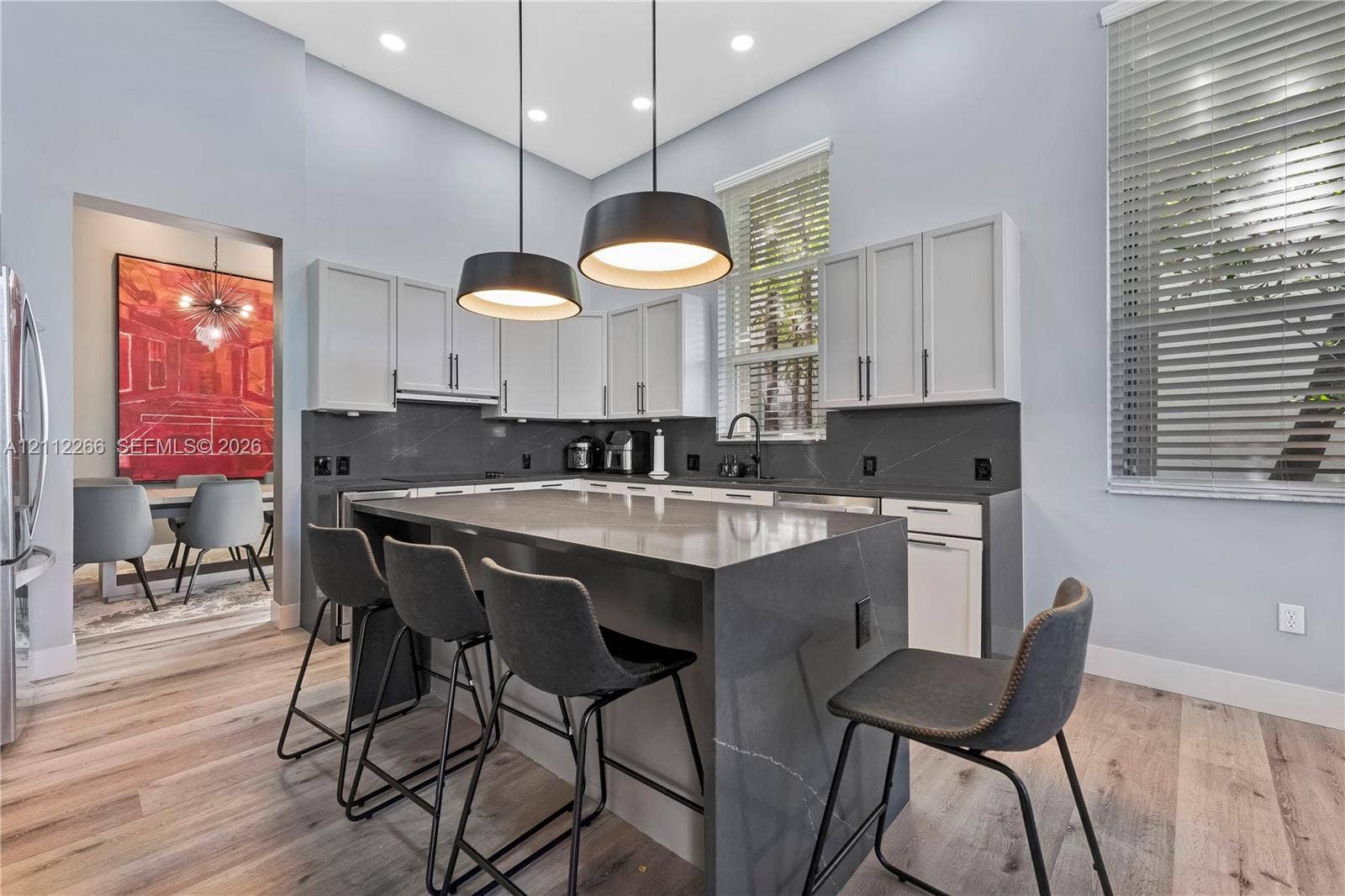 Kitchen featuring a central island with a solid-surface waterfall countertop, white cabinetry with dark hardware, dark solid-surface backsplash, wood-finish flooring, and dual pendant lighting