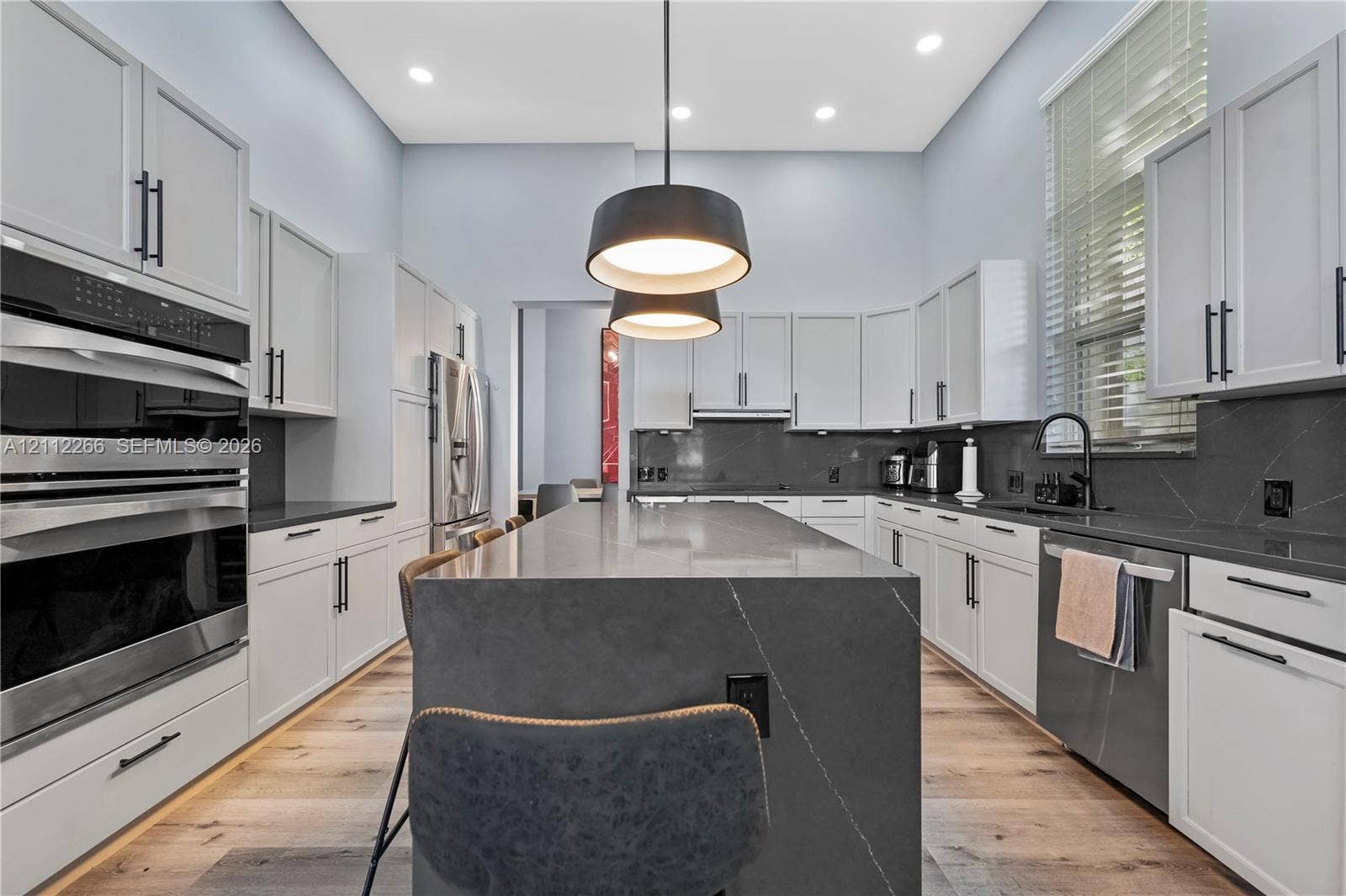 Kitchen featuring white shaker-style cabinetry, dark stone countertops with a waterfall-edge island, wood-finish flooring, dual wall ovens, and matte black hardware