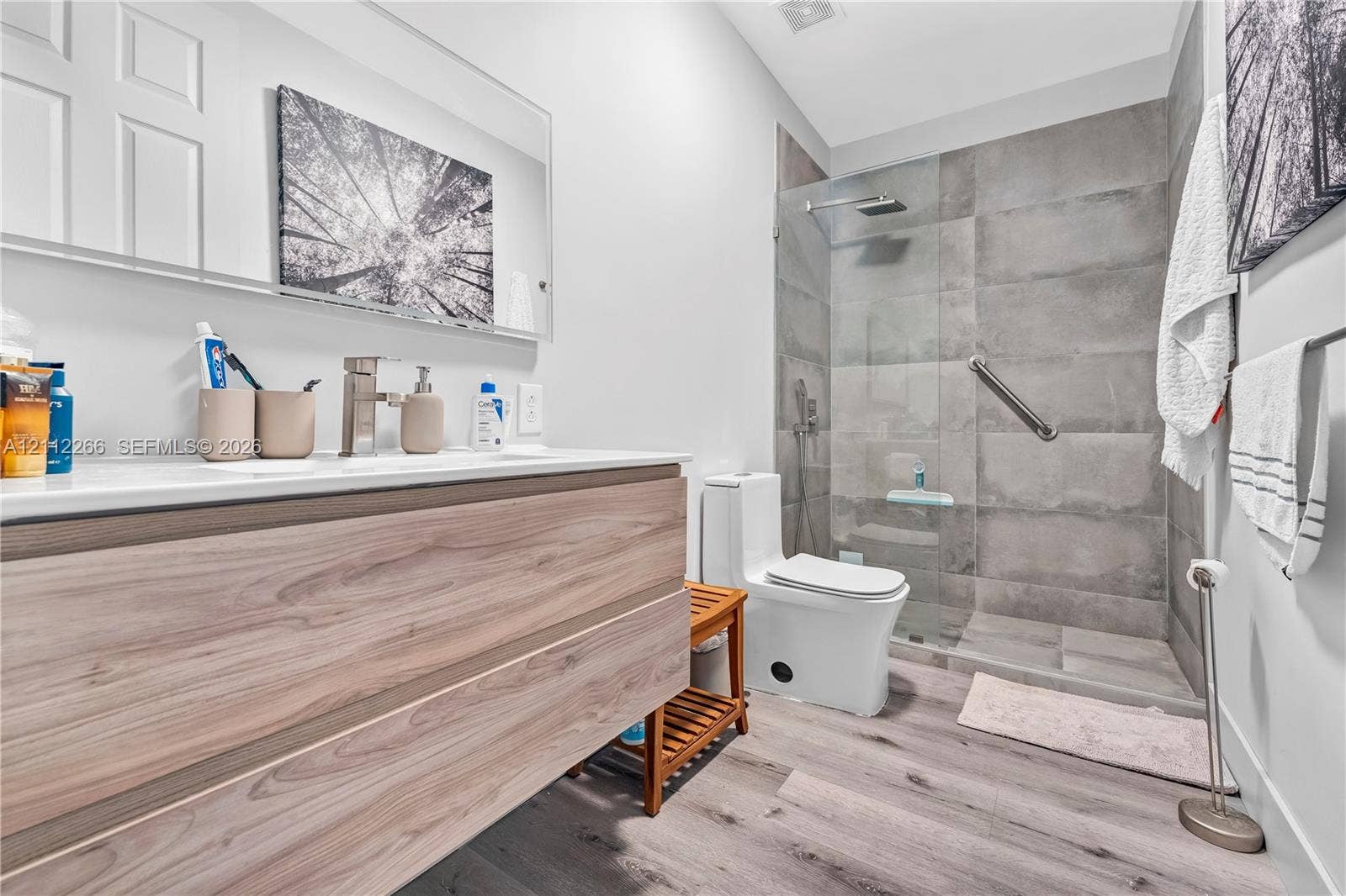 Floating wood-finish vanity with a clean-lined countertop, walk-in shower featuring large-format grey wall tile and a glass partition, rainfall showerhead, and wood-finish flooring