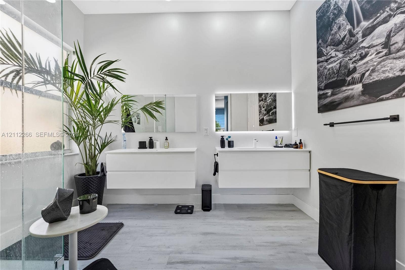 Dual white floating vanities featuring integrated sinks and minimalist cabinetry, paired with rectangular backlit mirrors, glass-enclosed shower, and wood-finish flooring