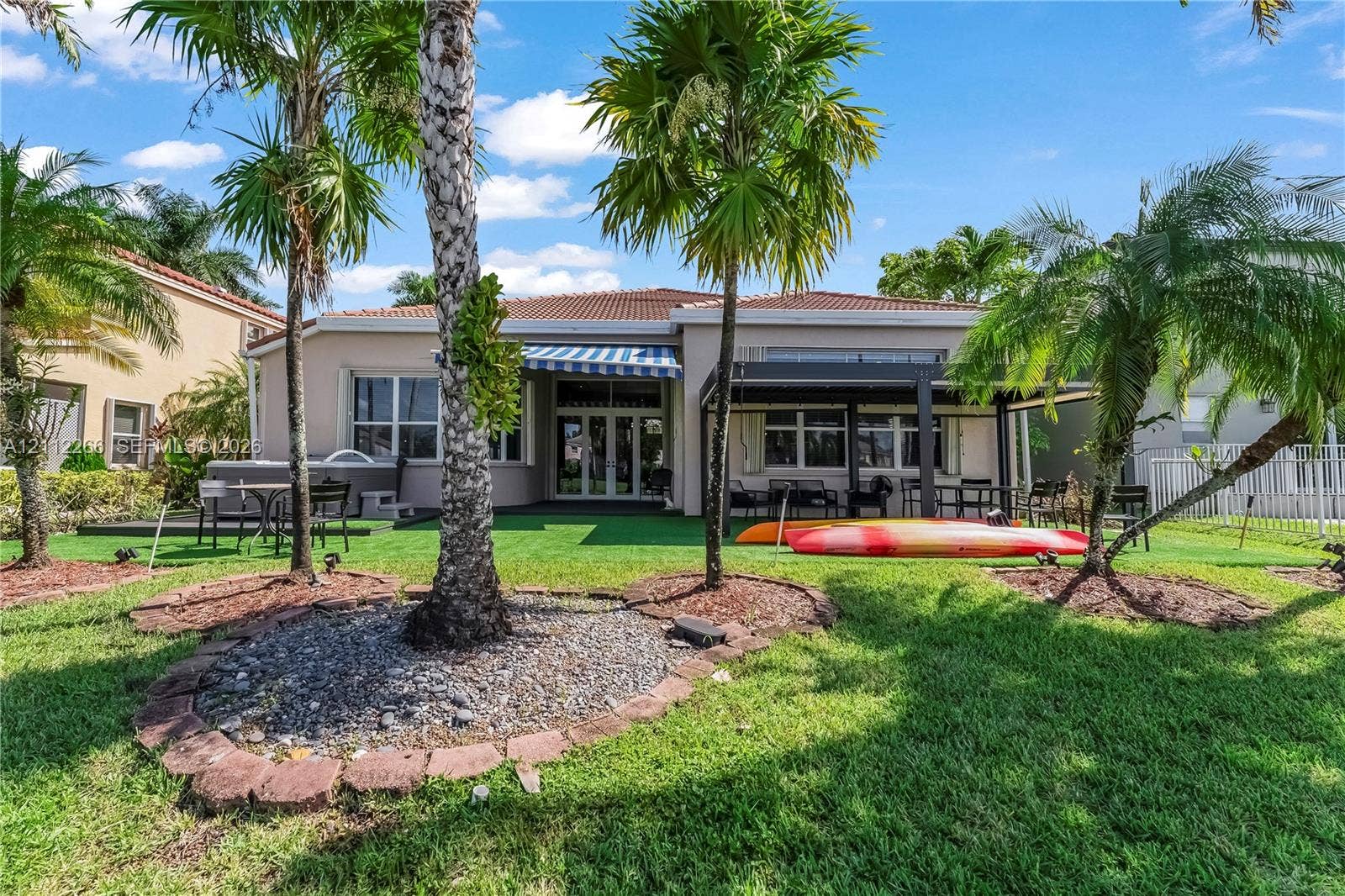 Backyard featuring a covered pergola, artificial turf, stone-bordered landscaping, mature palm trees, and an exterior striped awning