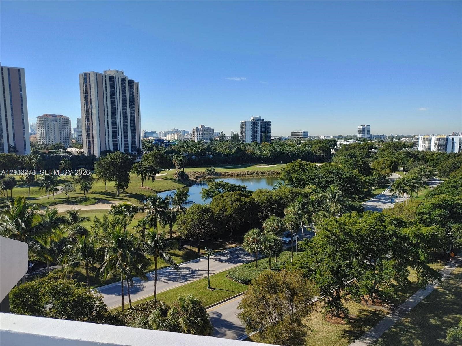 Elevated balcony perspective featuring expansive scenery of a manicured golf course, a central water feature, mature trees, and high-rise urban architecture
